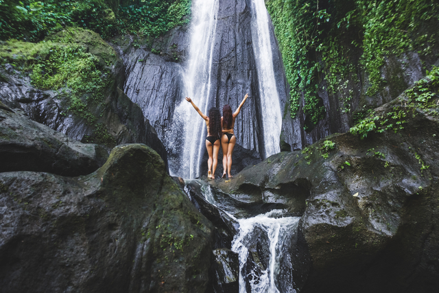 Two slim women relaxing near beautiful waterfall in Bali jungle. Nature adventure Dusun Kuning in Ubud area